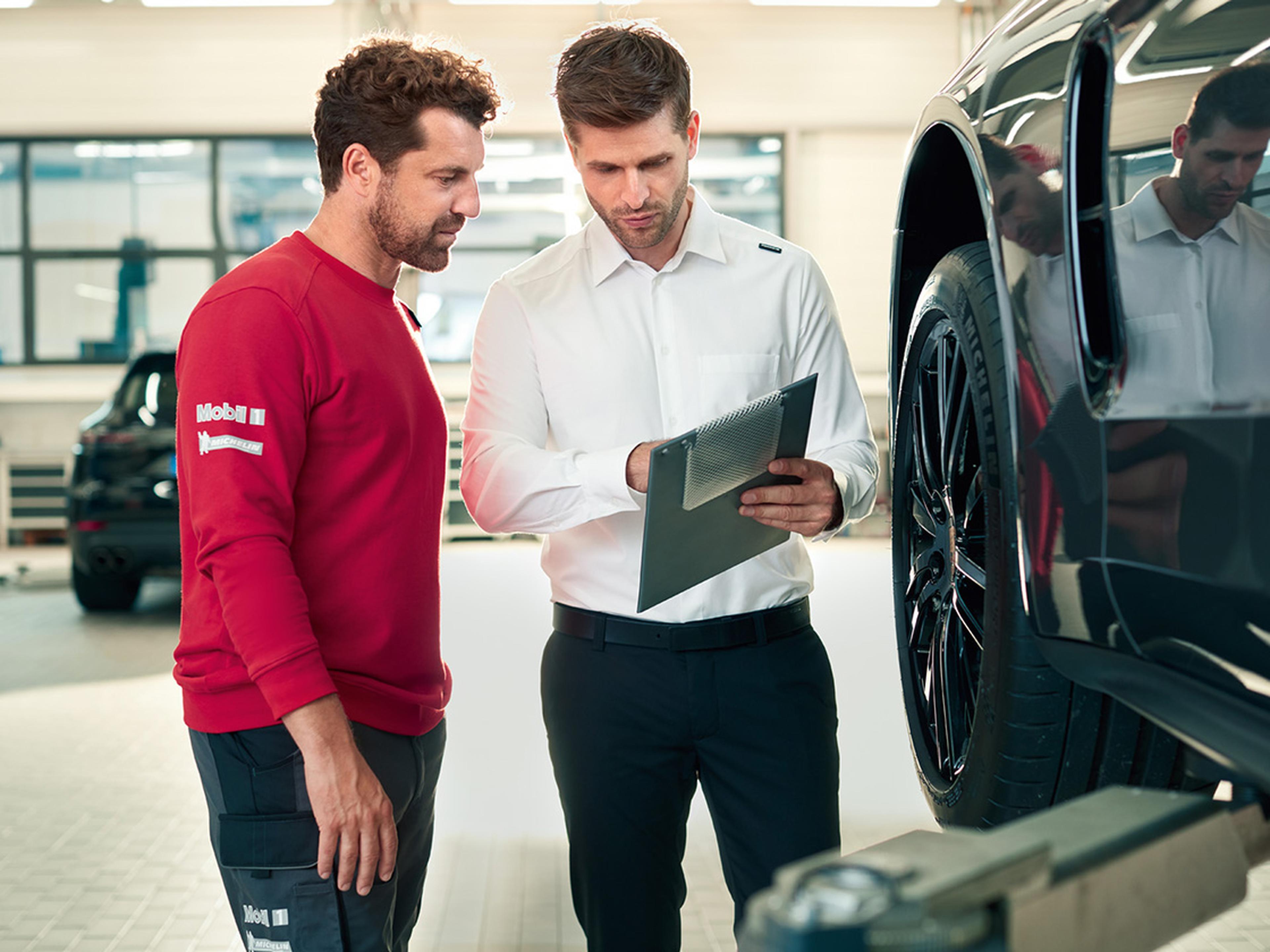 Zwei Porsche Mitarbeiter besprechen sich an der Frontachse eines Fahrzeugs. Einer hält ein Klemmbrett in Händen.