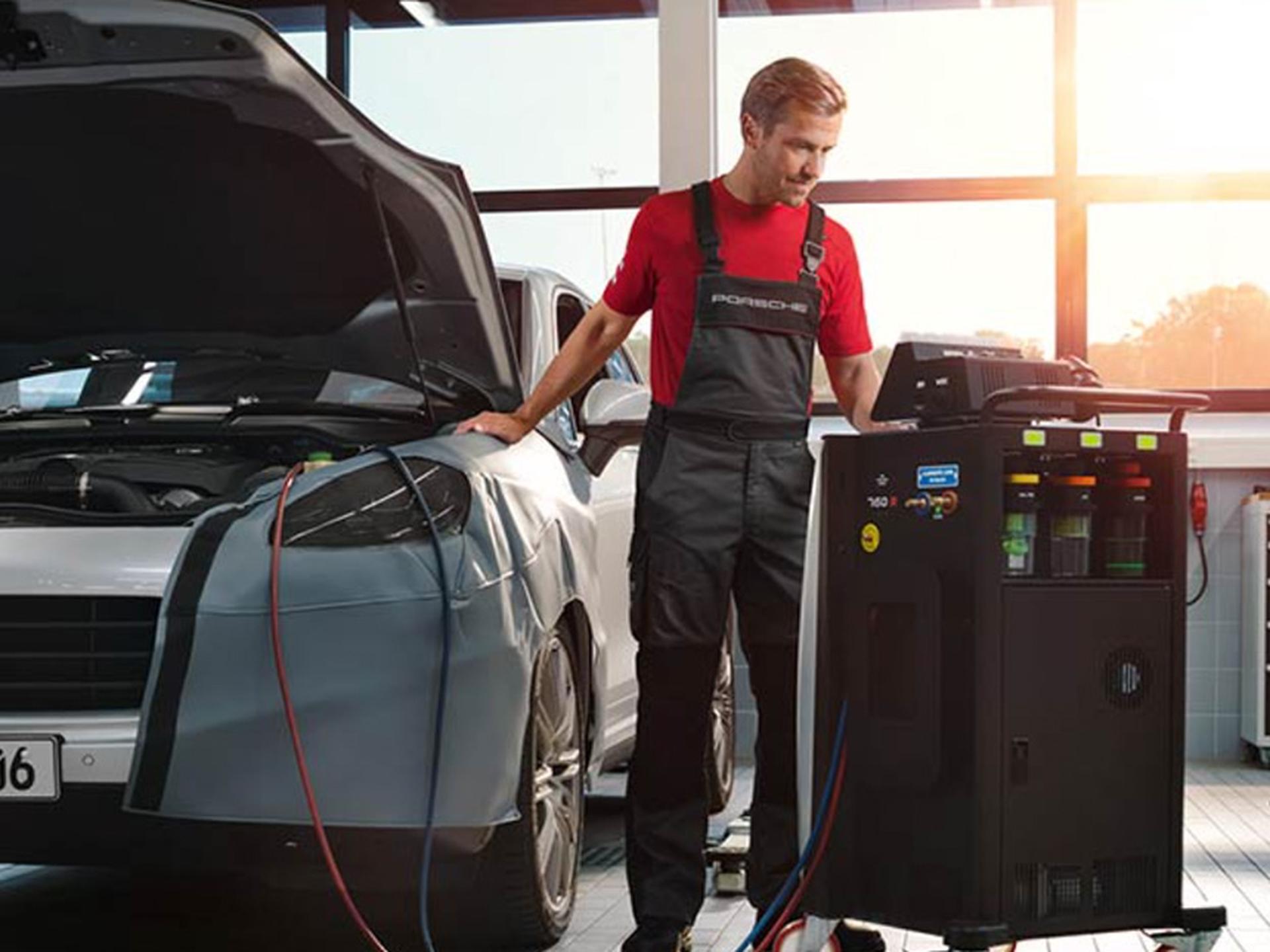 Porsche Mitarbeiter überprüft die Klimaanlage eines Fahrzeugs, das mit offener Motorhaube in der Werkstatt steht.