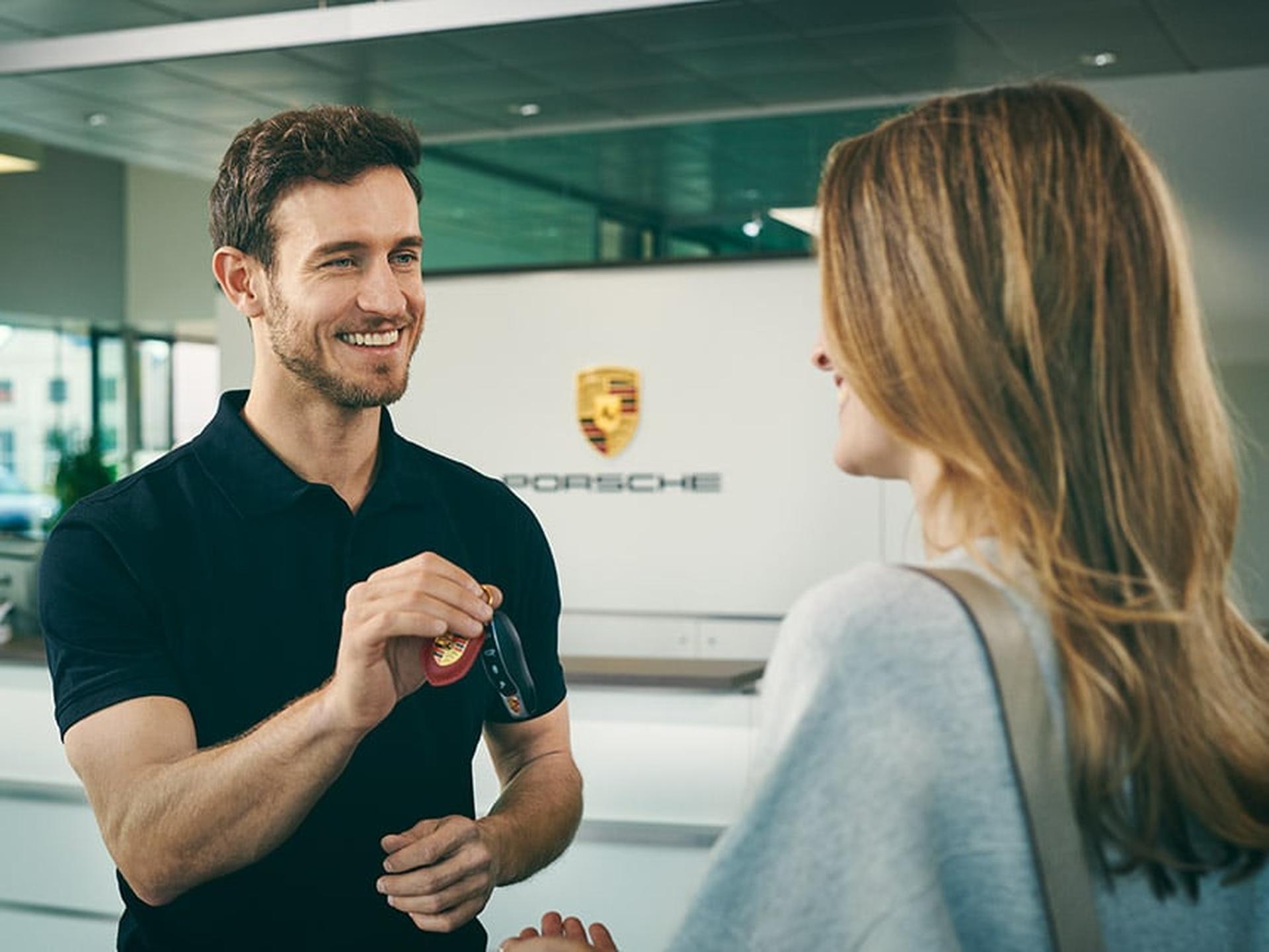 Porsche Mitarbeiter übergibt Porsche Drive Kundin mit einem Lächeln die Schlüssel für ihr Mietfahrzeug.