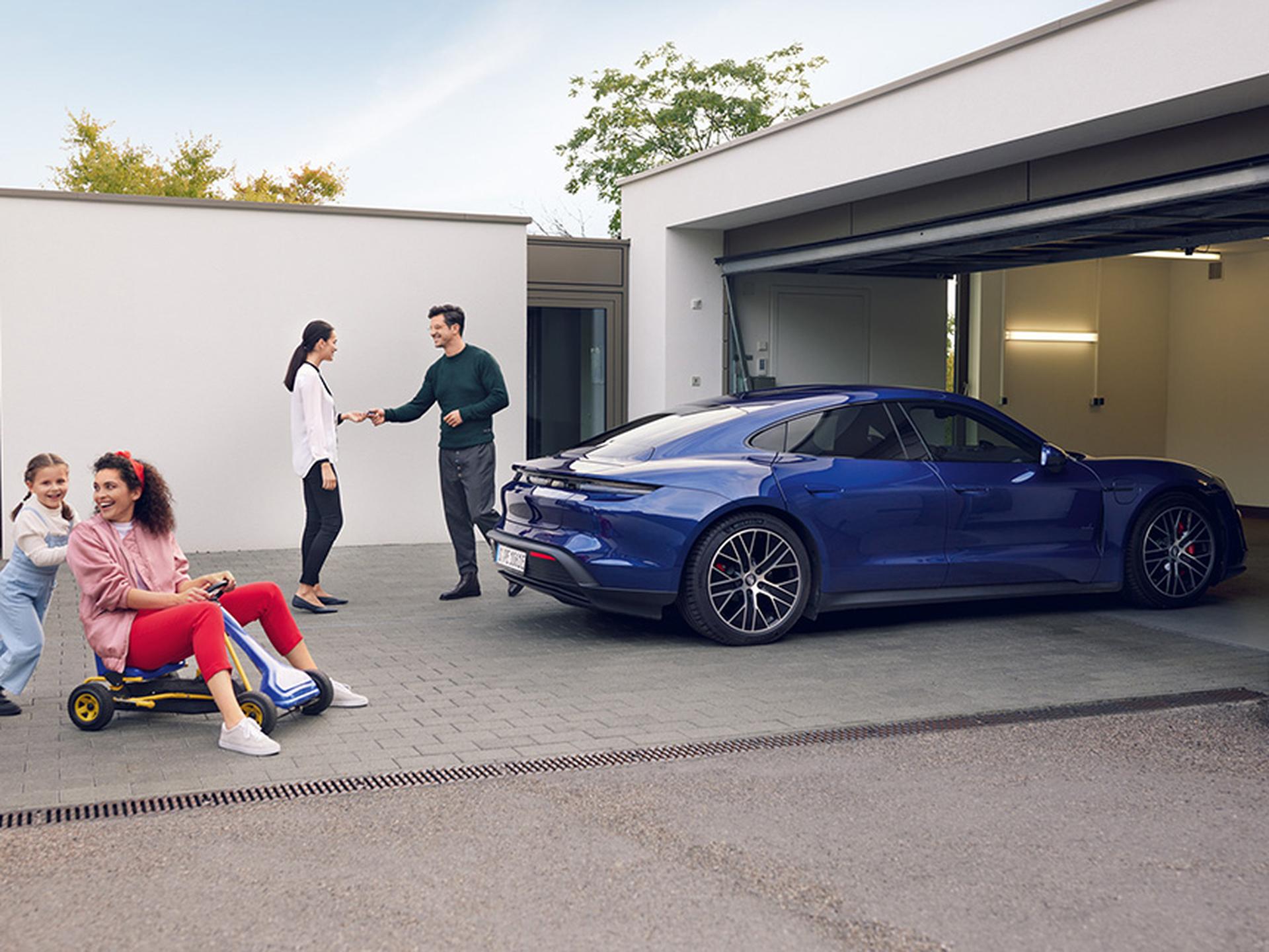 Porsche Mitarbeiter holt ein Fahrzeug bei einer Kundin ab. Kinder spielen im Hof.