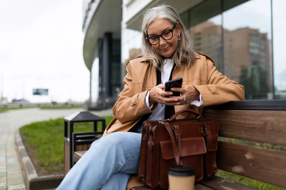 Frau nutzt ihr Smartphone auf einer Bank vor einem Bürogebäude.