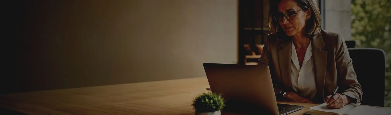 Eine Business-Frau sitzt an einem Schreibtisch vor ihrem Laptop und macht sich Notizen in einem Notizbuch.