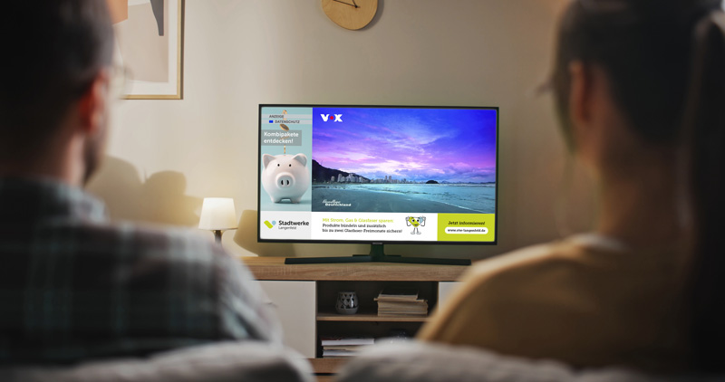 Ein Paar sitzt auf dem Sofa und über ihre Schultern kann man ein laufendes Fernsehprogramm mit einer ATV-L-Bannerwerbung sehen.