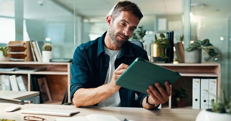 Ein Mann am Schreibtisch in seinem Büro hält ein Tablet in der Hand und arbeitet daran für sein Business.