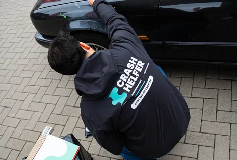 Gutachter prüft den Kotflügel eines schwarzen BMW, daneben Koffer und Gutachtenmappe
