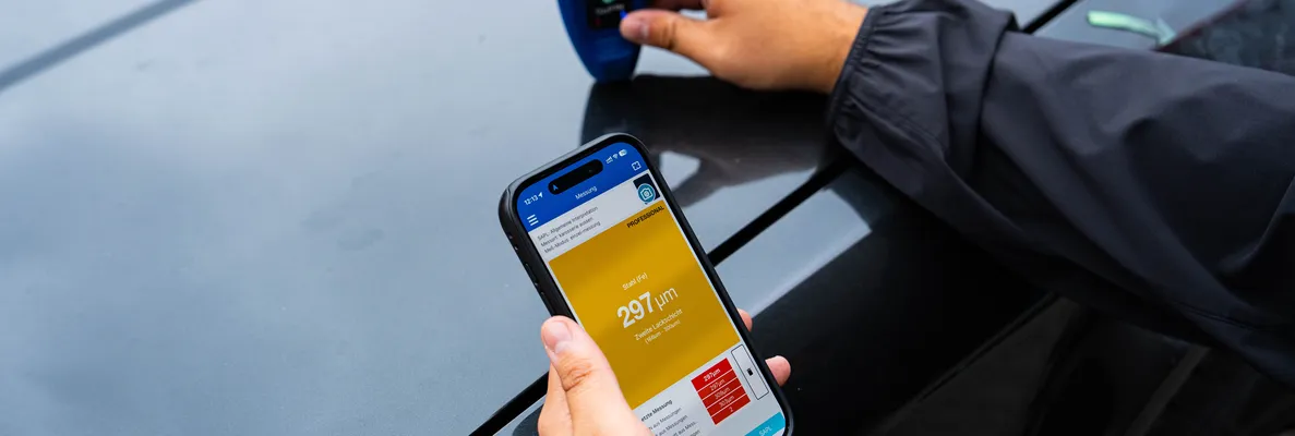 Gutachter misst die Lackschichtdicke an einem schwarzen Auto, der Messwert erscheint auf dem Smartphone
