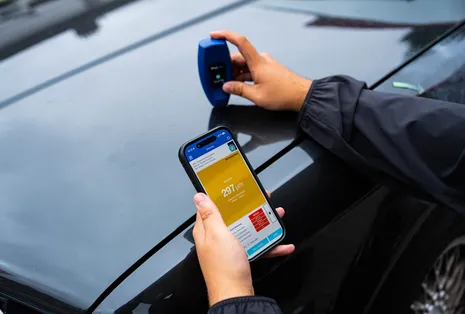 Gutachter misst die Lackschichtdicke an einem schwarzen Auto, der Messwert erscheint auf dem Smartphone