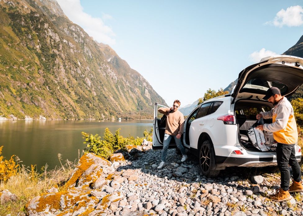 Two men by a white SUV with open trunk beside a scenic fjord surrounded by mountains on a sunny day.