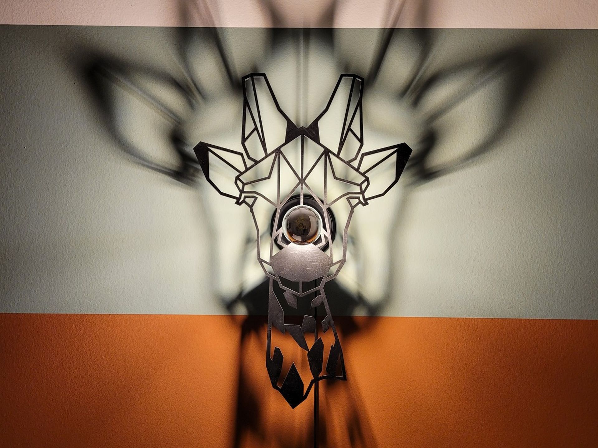 Detail van de geometrische giraffelamp met schaduwspel op een roestbruine en groengrijze wand.