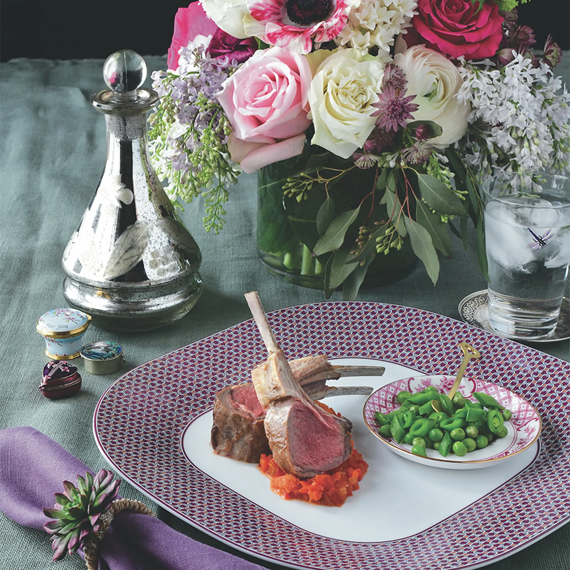 A plated rack of lamb with sautéed peas and vegetables on a table with flowers