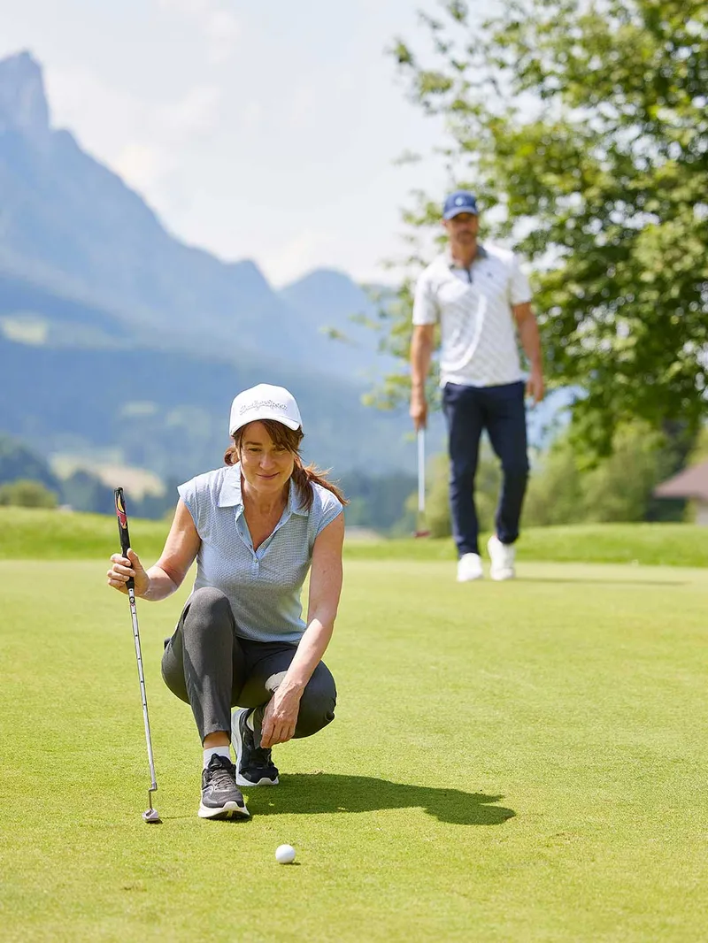 Golferin kniet zur Ballanalyse auf dem Grün, im Hintergrund Berge und Mitspieler