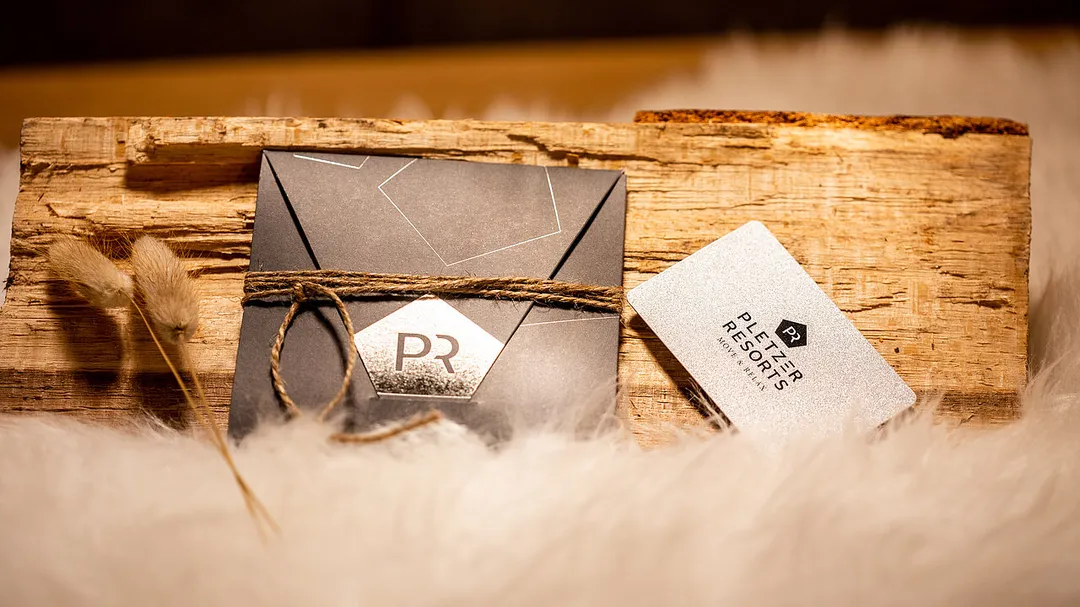 Ein dunkler Umschlag mit "PR"-Logo und eine glitzernde Pletzer Resorts Karte auf rustikalem Holz und weißem Fell, mit Trockenblumen.