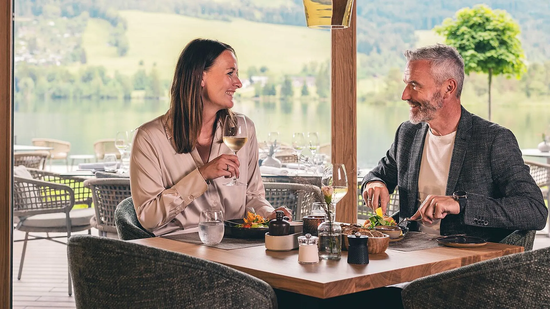 Ein lächelndes Paar genießt ein Essen mit Wein in einem Restaurant mit Seeblick und grüner Landschaft im Hintergrund.