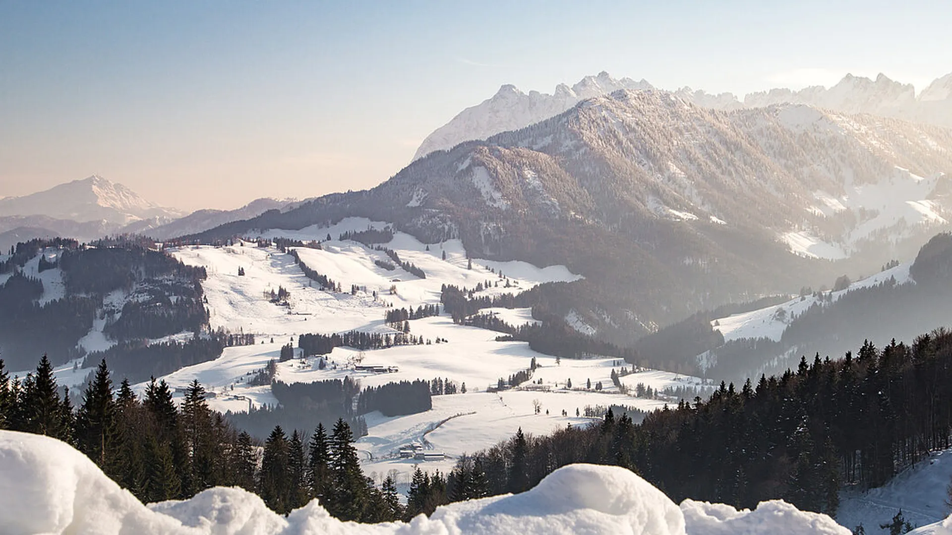 Weite, verschneite Berglandschaft mit Nadelwäldern, hell erleuchteten Tälern und hohen, sonnenverwöhnten Gipfeln.