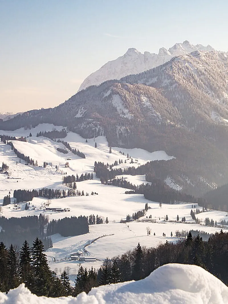 Weite, verschneite Berglandschaft mit Nadelwäldern, hell erleuchteten Tälern und hohen, sonnenverwöhnten Gipfeln.