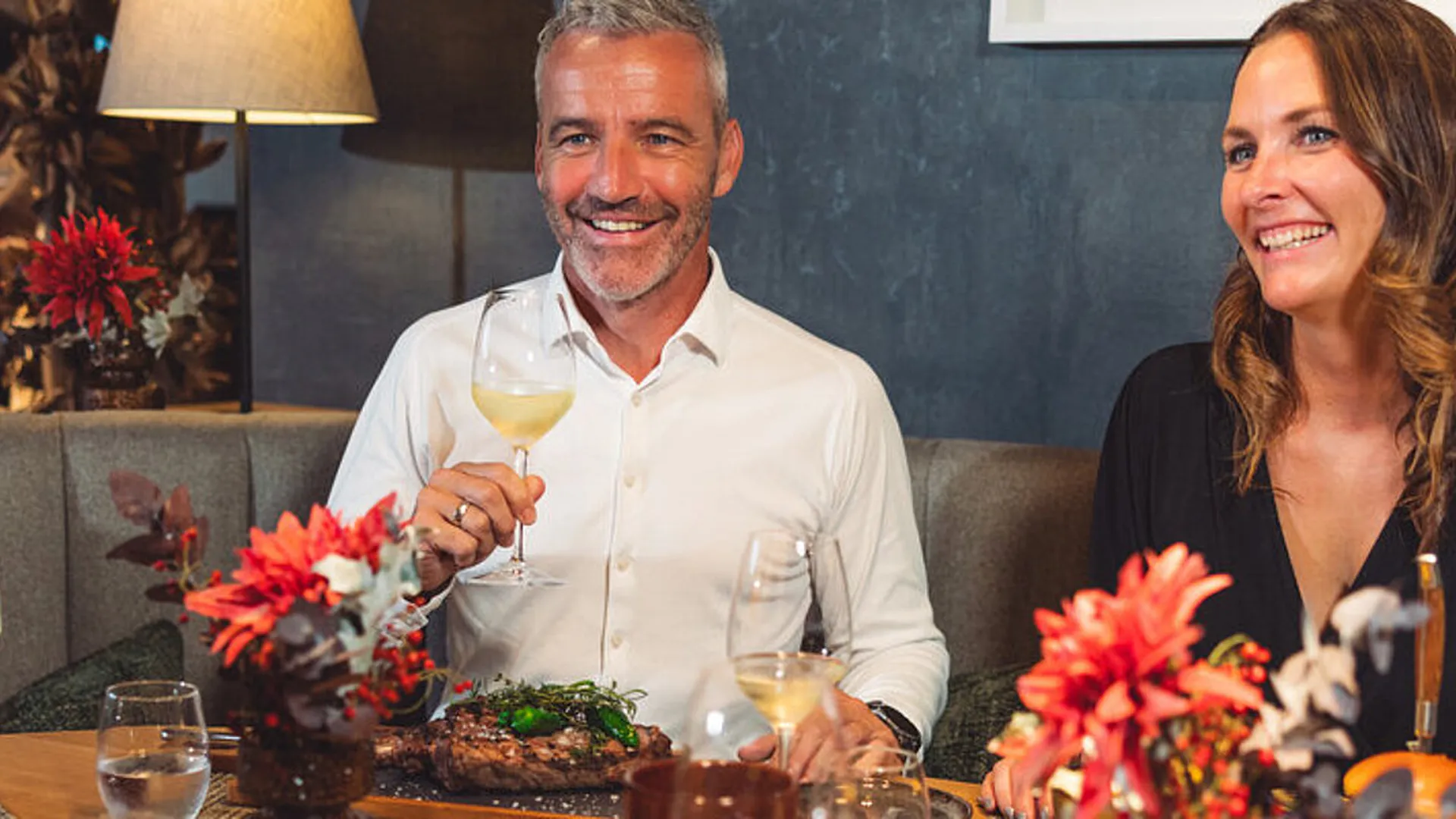 Lächelndes Paar beim Abendessen im Restaurant mit Wein und Steak. Rote Blumen schmücken den Tisch.