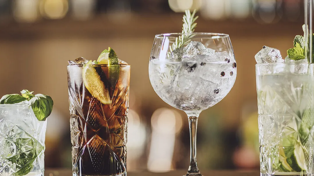 Vier verschiedene Cocktails auf einer Bar: Ein Basilikum-Drink, Cuba Libre, Gin Tonic mit Rosmarin und ein Mojito, alle mit Eis und Garnierung.