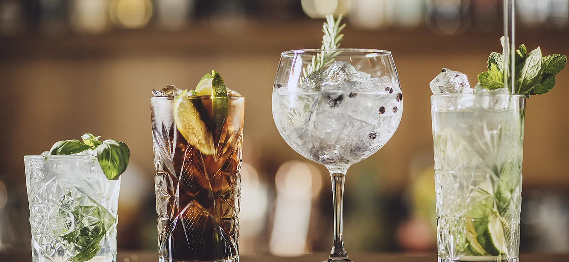 Vier verschiedene Cocktails auf einer Bar: Ein Basilikum-Drink, Cuba Libre, Gin Tonic mit Rosmarin und ein Mojito, alle mit Eis und Garnierung.