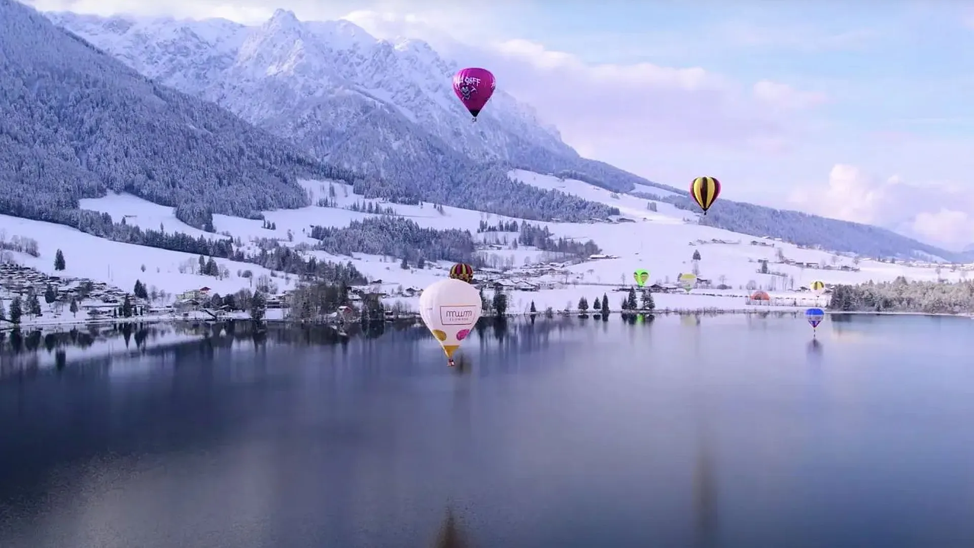 Mehrere bunte Heißluftballons schweben über einem ruhigen See, der eine verschneite Berglandschaft und ein Dorf am Ufer reflektiert.