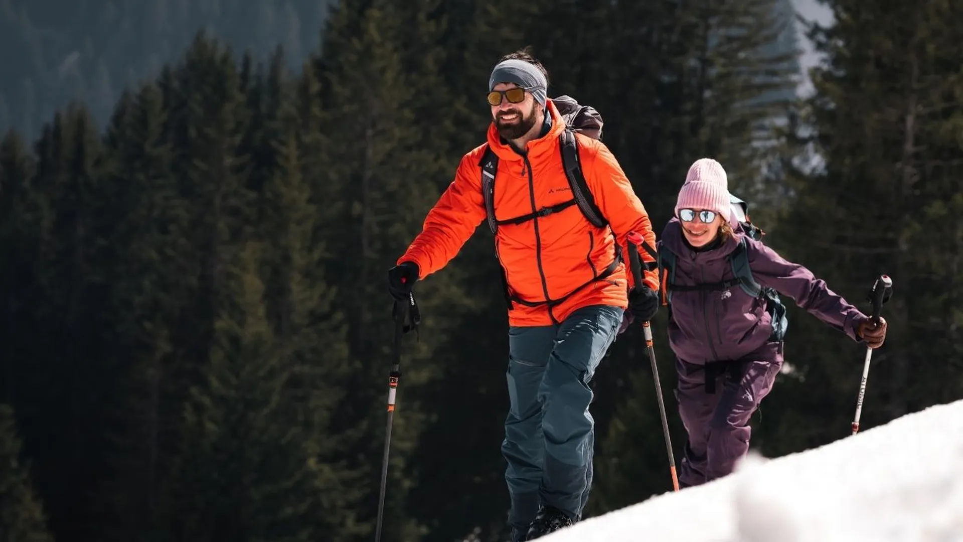 Zwei Personen mit Skiern und Stöcken bei einer Skitour im Schnee vor Nadelwald