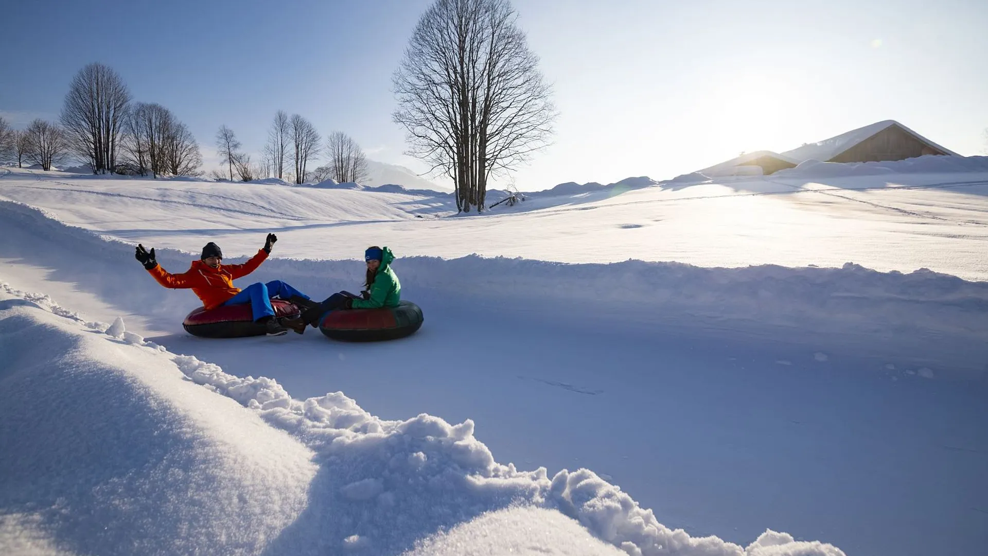 Zwei Personen rodeln auf Reifen eine verschneite Bahn hinunter. Sonnenschein erhellt die Winterlandschaft mit Bäumen im Hintergrund.