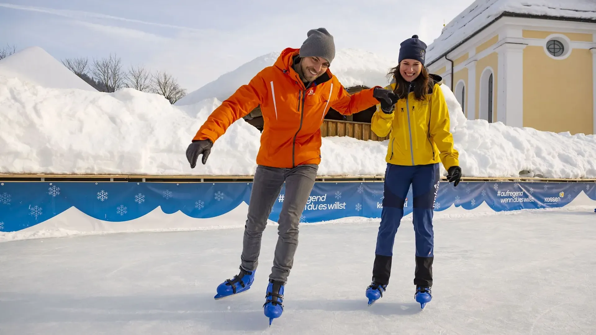 Ein lächelndes Paar beim Eislaufen auf einer Eisbahn im Winter, hält Händchen. Im Hintergrund viel Schnee und ein Gebäude.