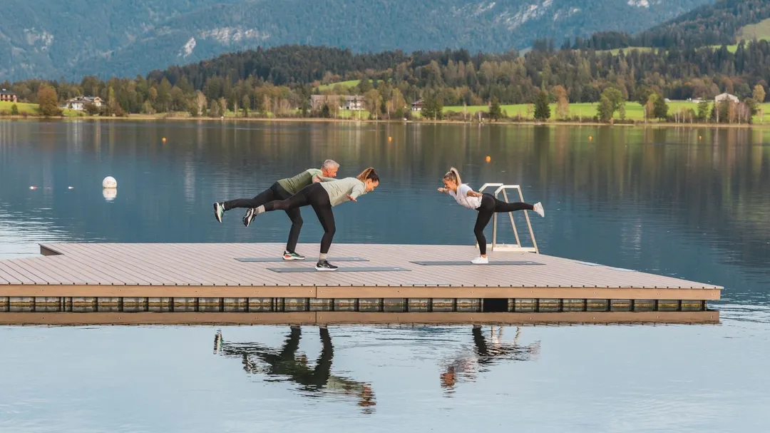 Personen machen Yoga auf einem Steg am See vor Bergpanorama