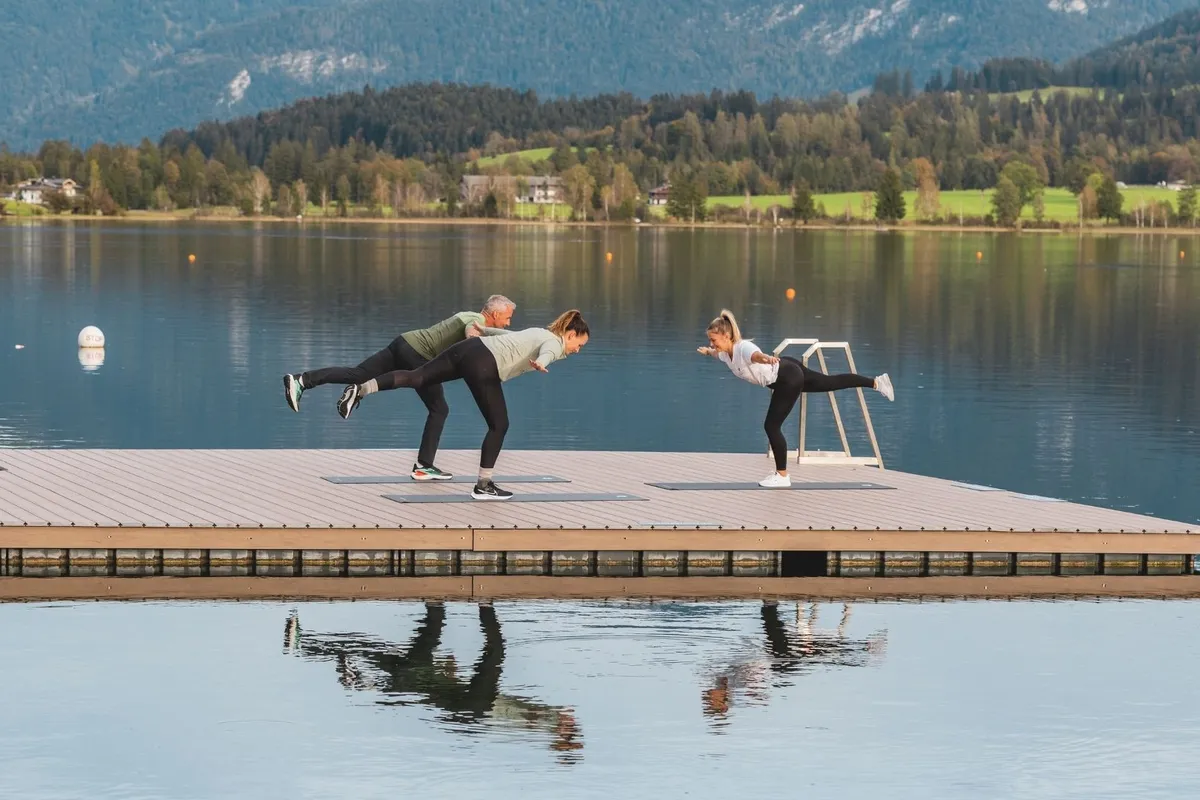 Personen machen Yoga auf einem Steg am See vor Bergpanorama
