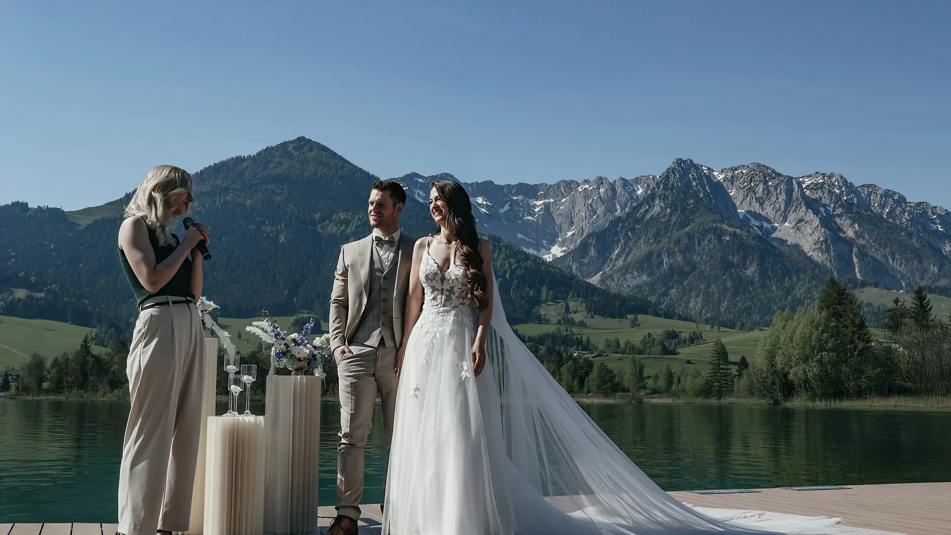 Trauung am See: Brautpaar und Traurednerin vor Alpenkulisse an sonnigem Tag.
