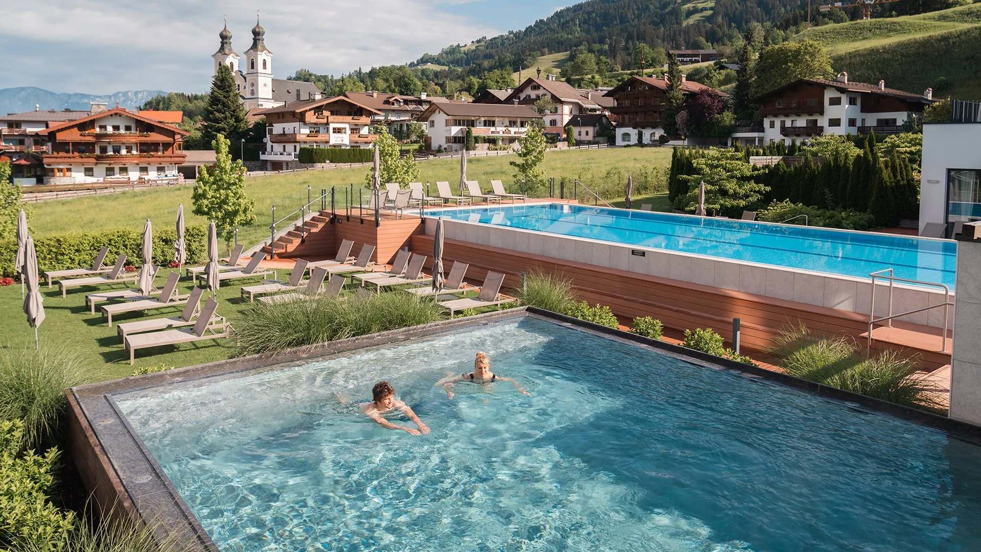Außenpools mit Liegestühlen, im Hintergrund Dorf mit Kirche und Berge