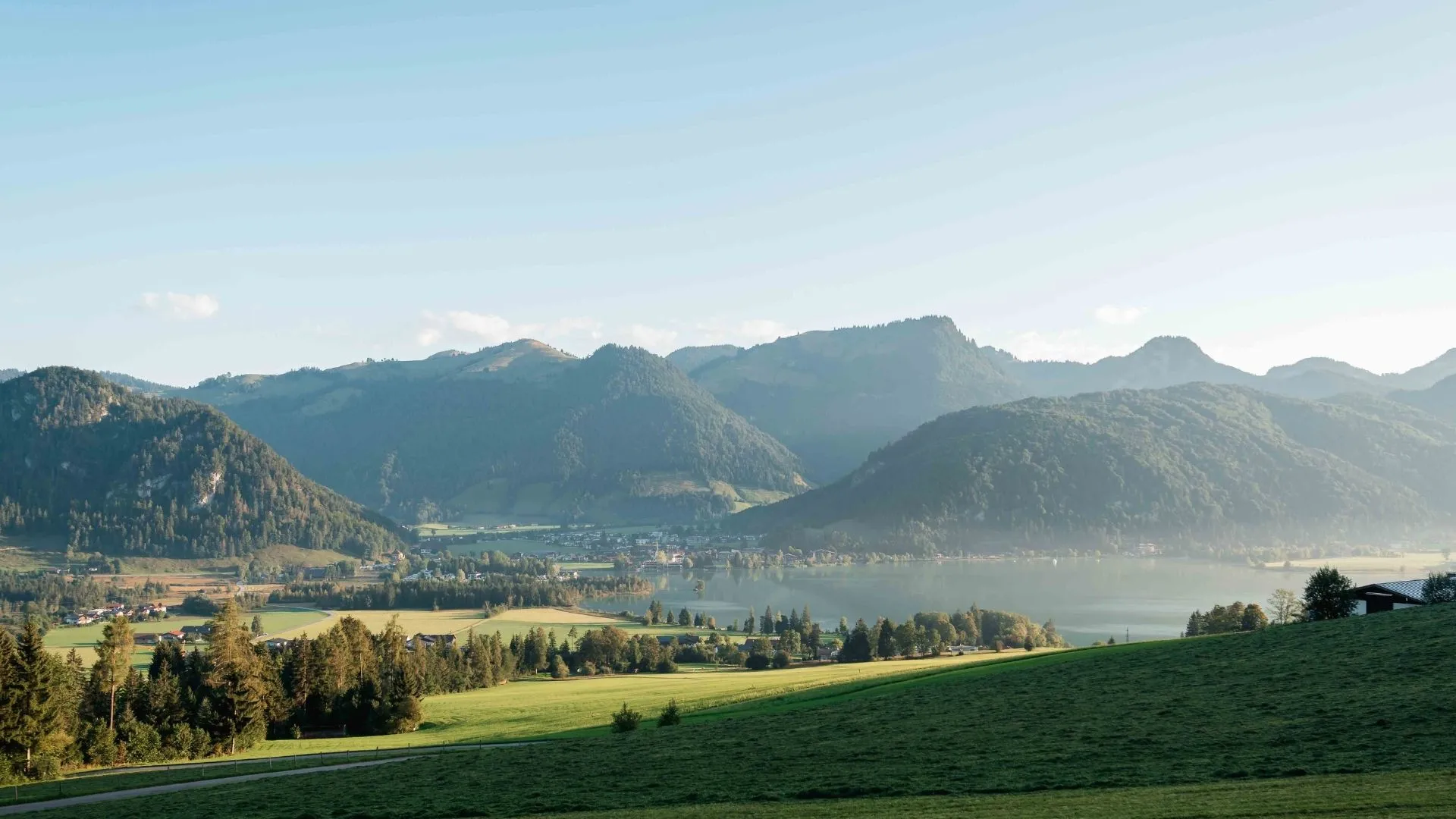 Blick über grüne Wiesen auf einen Alpensee mit umliegenden Bergen