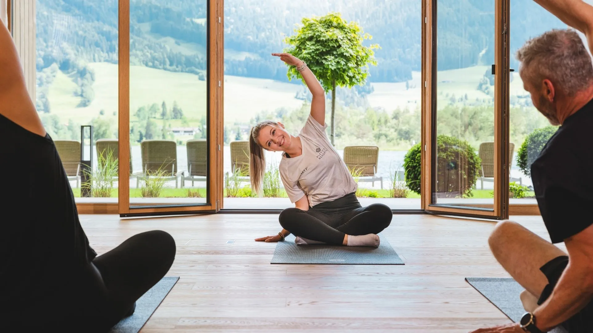 Yogastunde mit Trainerin und Gästen in einem Raum mit Panoramafenstern und Bergblick