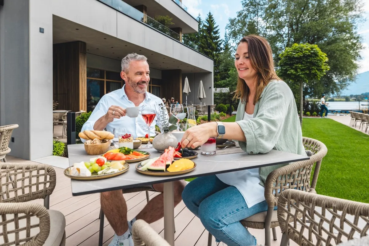 Paar frühstückt auf Terrasse am See mit Obstplatten und Getränken