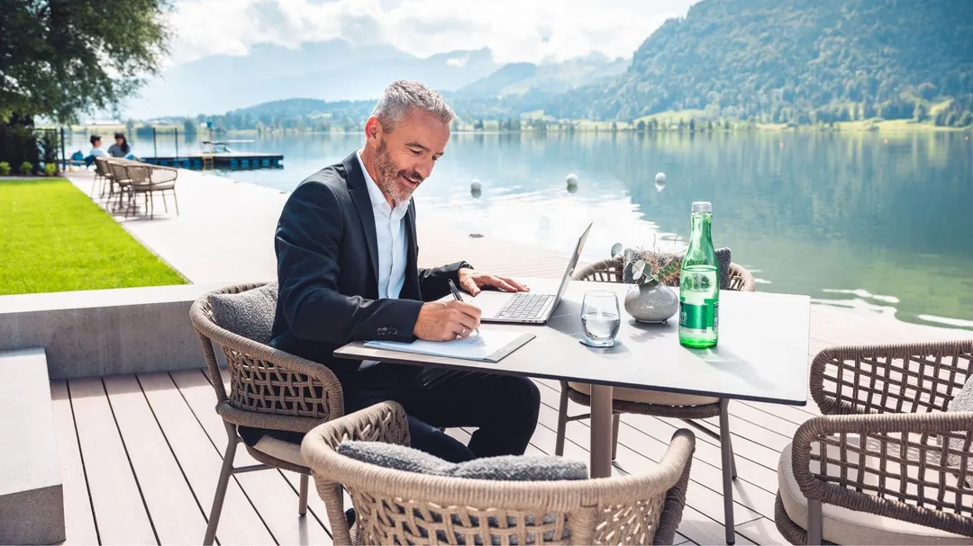 Mann arbeitet mit Laptop an einem Terrassentisch am See vor Bergkulisse