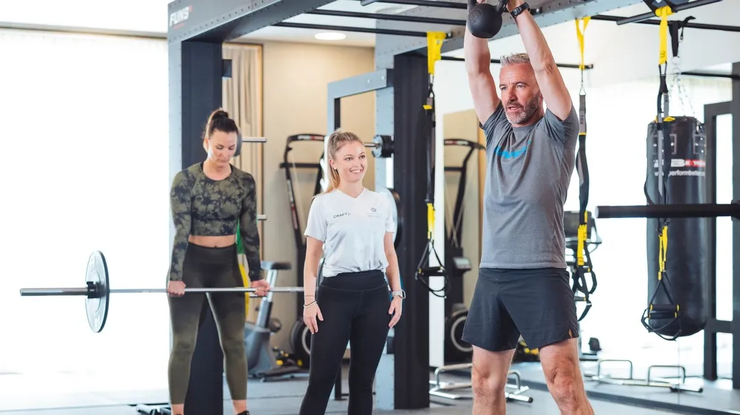 Trainer und Gäste beim Krafttraining mit Kettlebell und Langhantel im Fitnessraum