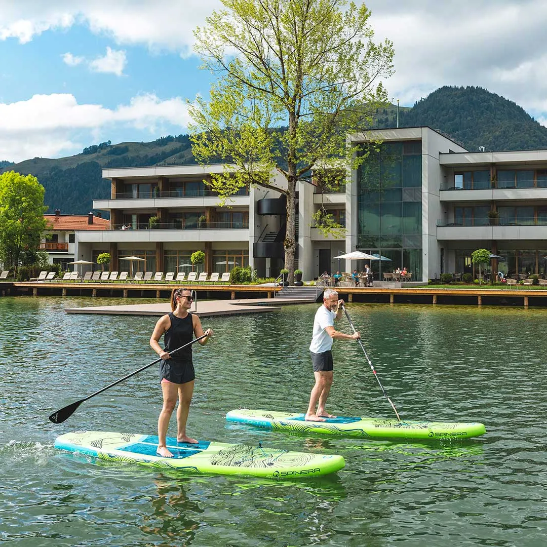 Paar beim Stand-Up-Paddling auf See vor Hotel mit Bergpanorama