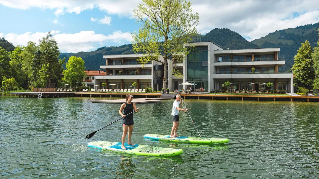 Paar beim Stand-Up-Paddling auf See vor Hotel mit Bergpanorama