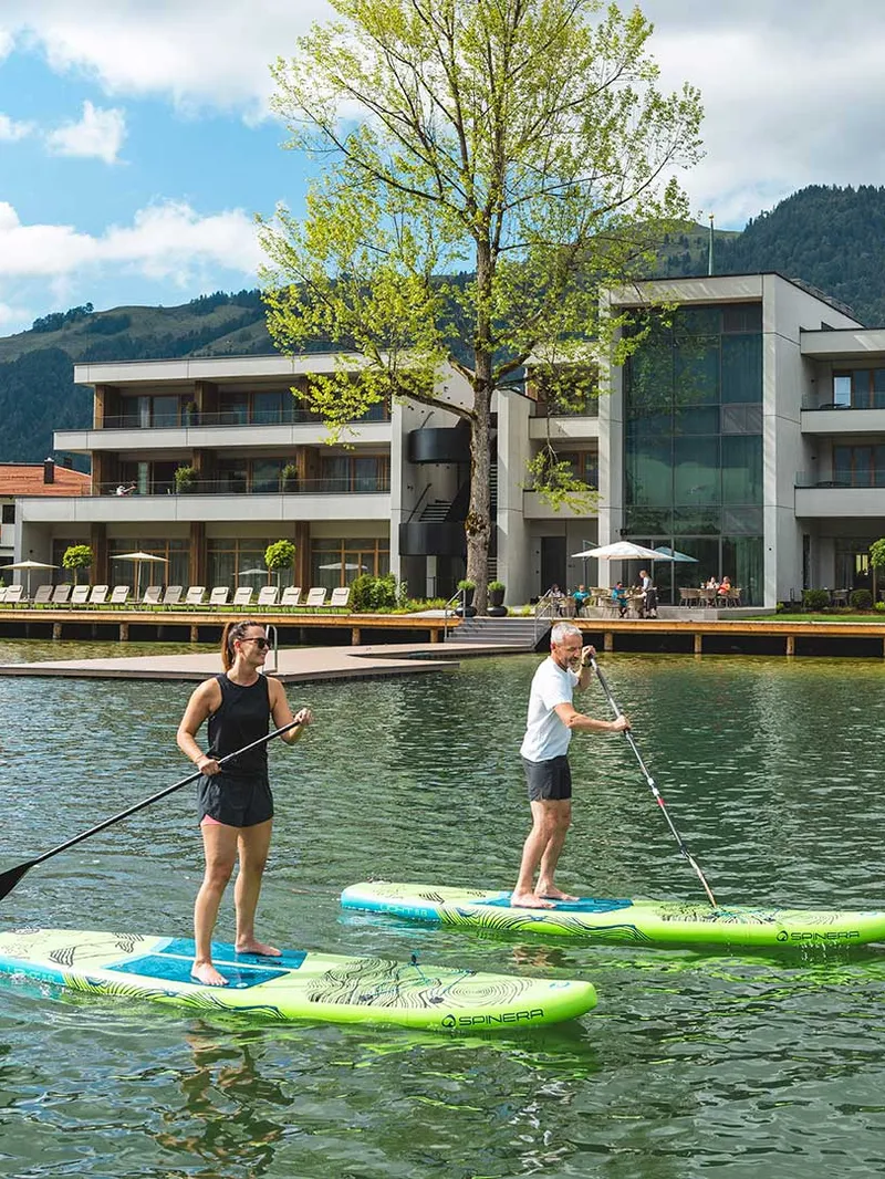 Paar beim Stand-Up-Paddling auf See vor Hotel mit Bergpanorama
