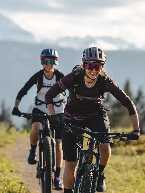 Zwei Frauen fahren mit Mountainbikes auf einem Bergpfad vor Alpenpanorama