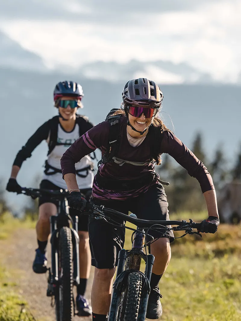 Zwei Frauen fahren mit Mountainbikes auf einem Bergpfad vor Alpenpanorama