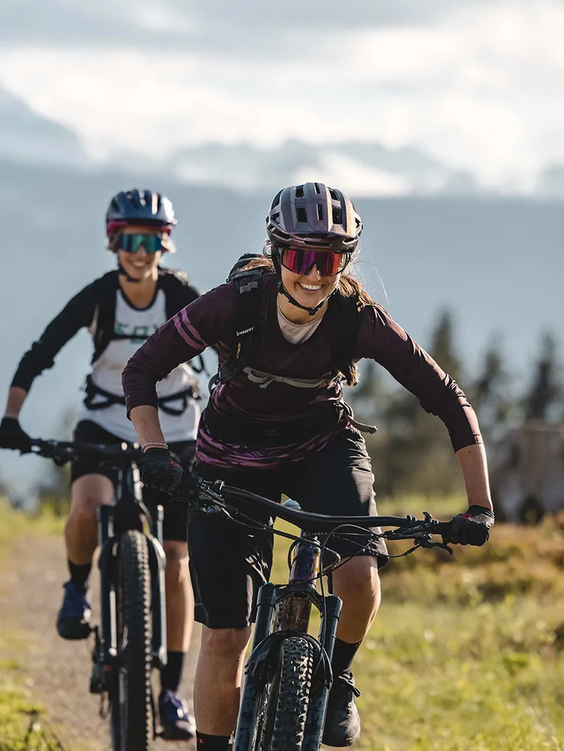 Zwei Frauen fahren mit Mountainbikes auf einem Bergpfad vor Alpenpanorama