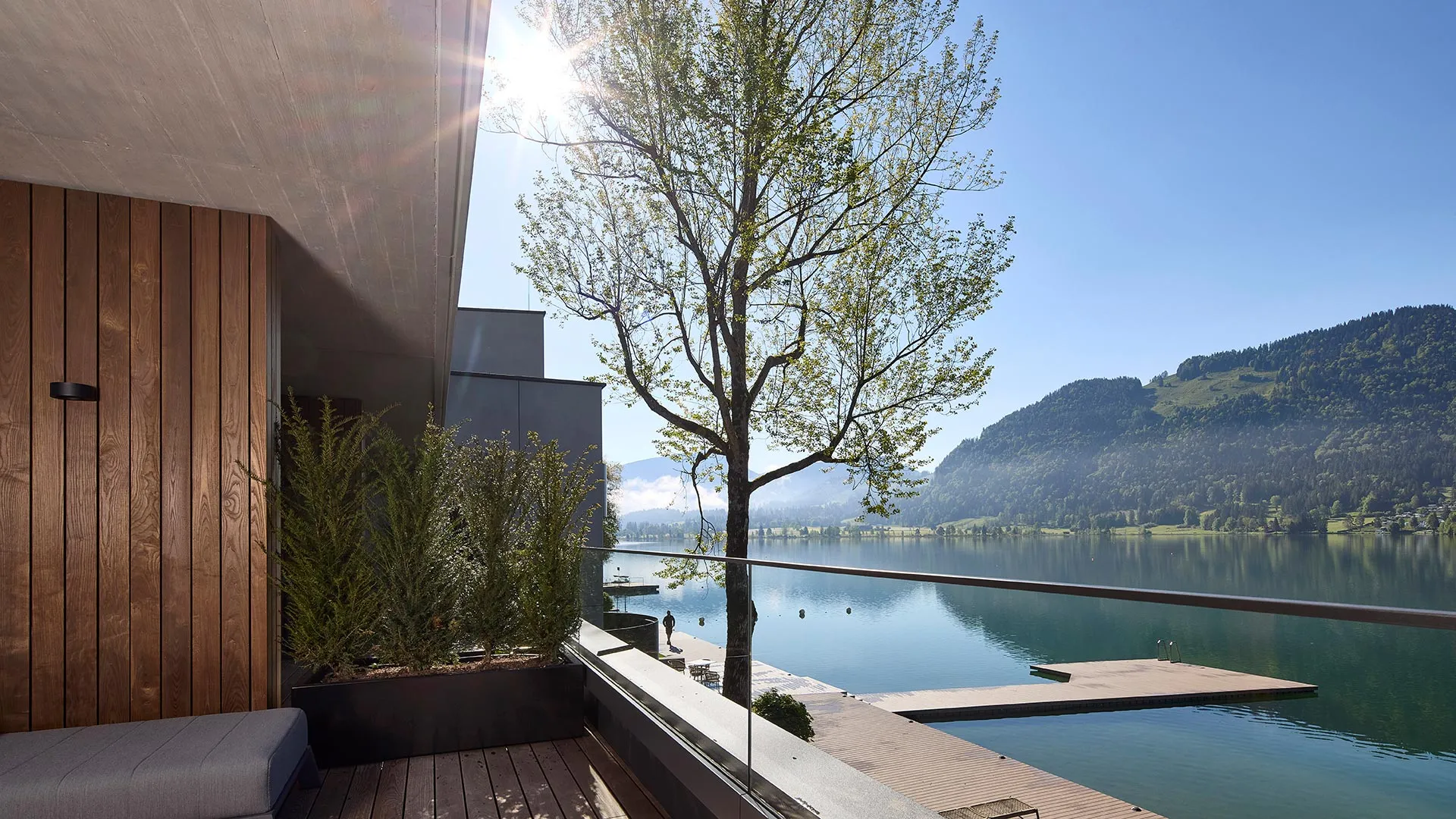 Moderner Balkon mit Holzboden und Glasgeländer, Blick auf einen ruhigen See mit bewaldeten Bergen, großem Baum und hölzernen Stegen.