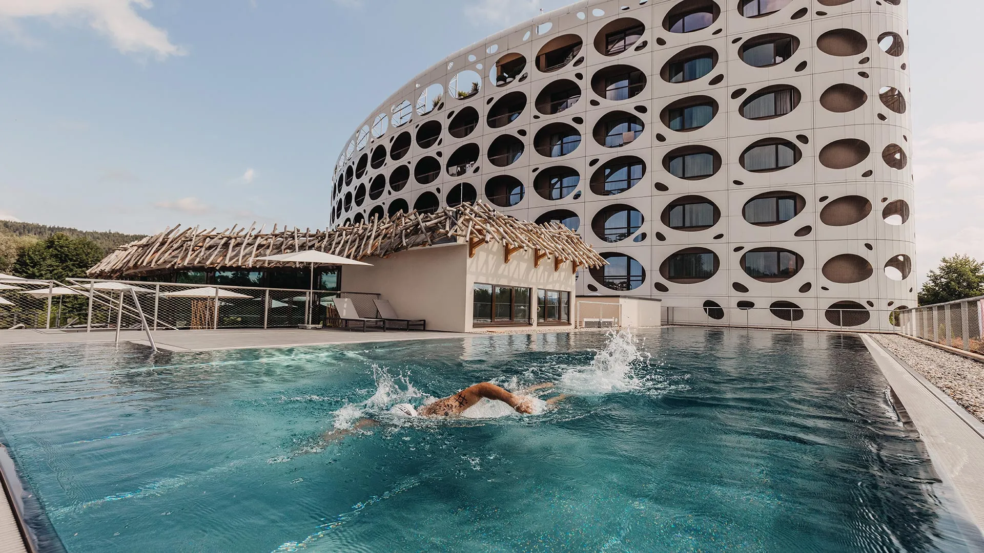 Schwimmer im Außenpool vor markanter Hotelfassade mit ovalen Fensteröffnungen