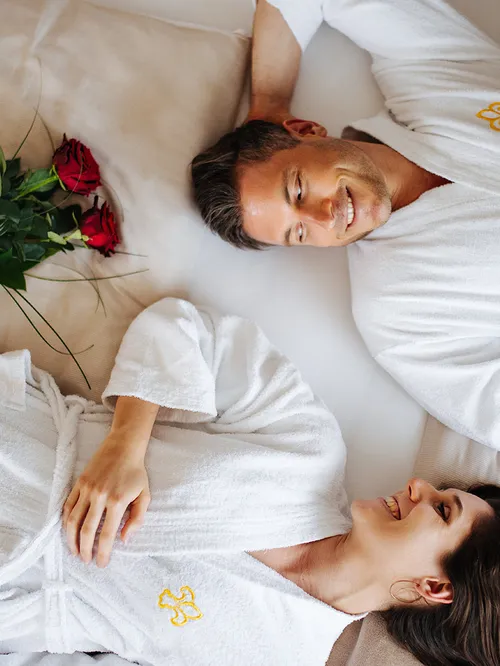 Paar in Bademänteln liegt lächelnd auf einem Bett mit roten Rosen