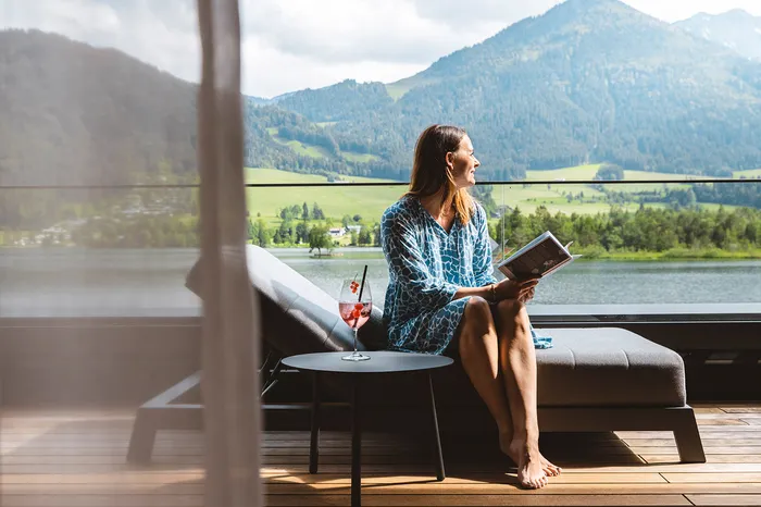 Entspannte Frau liest auf Balkon mit Cocktail, genießt den Panoramablick auf See und grüne Berge.