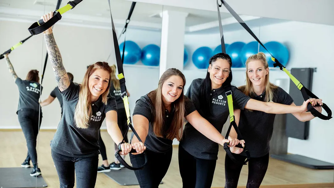 Vier lächelnde Mitarbeiterinnen der Pletzer Resorts trainieren mit TRX-Bändern im Fitnessraum