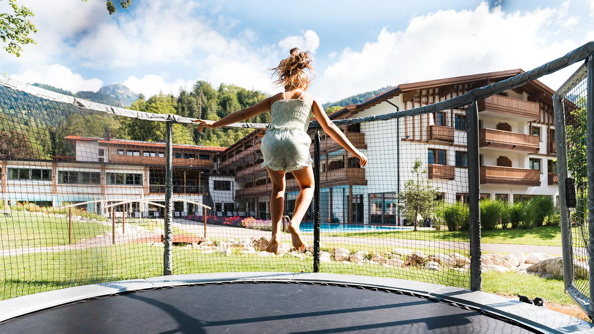 Frau springt auf Trampolin vor Hotelgebäude mit Pool und Bergen