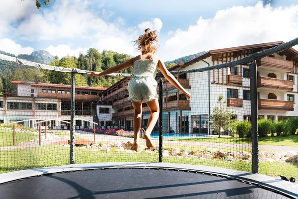 Frau springt auf Trampolin vor Hotelgebäude mit Pool und Bergen