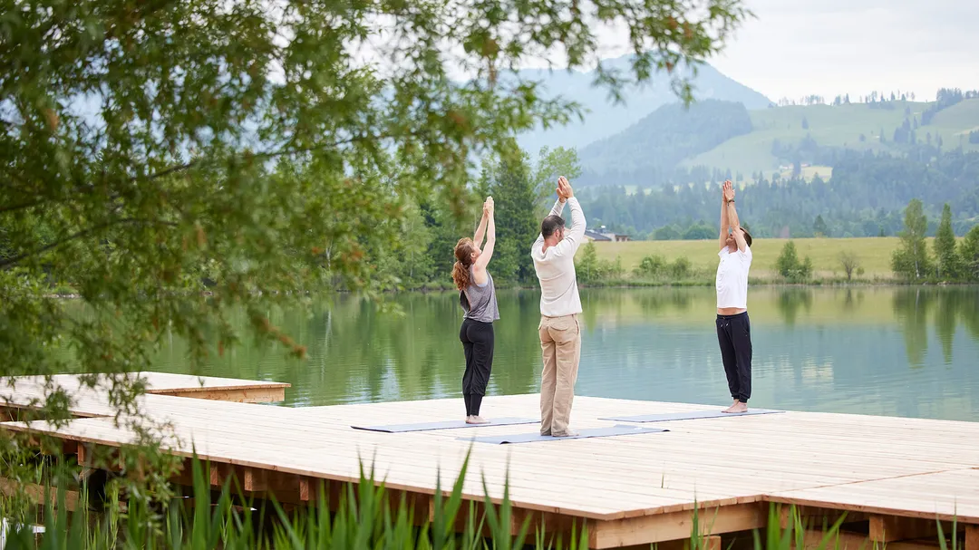 Personen praktizieren Yoga auf einem Holzsteg am See vor Bergkulisse