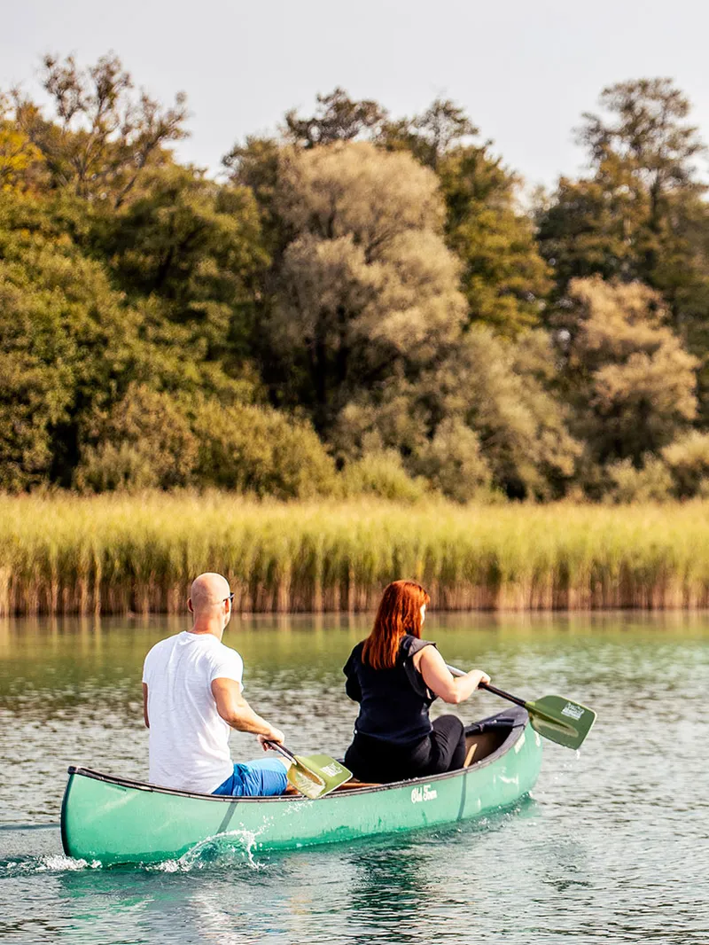 Paar paddelt in einem grünen Kanu auf einem See vor Schilf und Bäumen